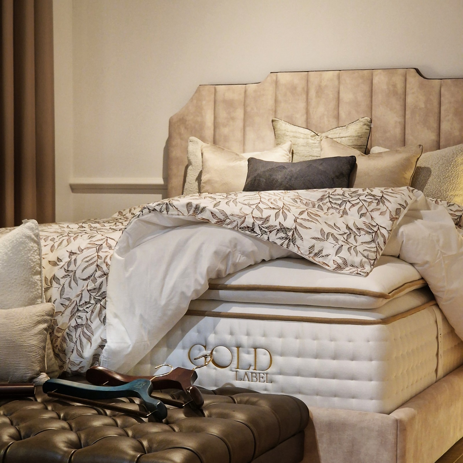 Elegant bed with a plush headboard, adorned with decorative pillows and a patterned comforter, featuring a 'GOLD LABEL' mattress. A wooden hanger rests on a tufted ottoman in the foreground.