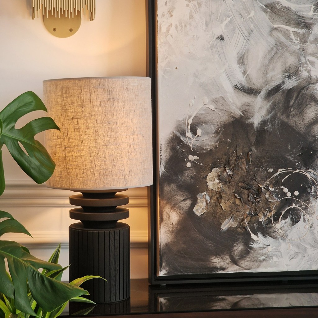 A modern table lamp with a grey fabric shade is placed next to a framed abstract painting featuring dark and light swirls with textures, alongside a large green leaf plant.